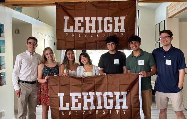A group of students at the Seattle Sendoff stand together and hold a brown and white Lehigh banner. A Lehigh flag is behind them.