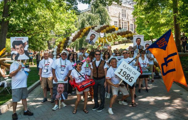 Members of the Class of 1986 smile and hold up signs with characters from Ferris Bueller's Day Off as they wave their class flag. 
