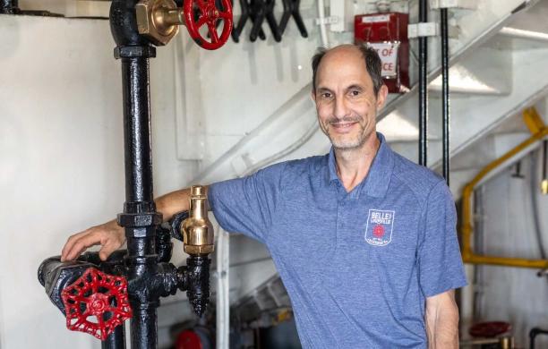 Tom stands in the engine room of the Belle with an arm draped over several pipes. He wears a blue shirt with the Belle logo on it.