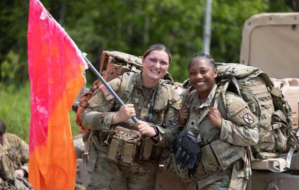 Maura wears her military fatigues and poses with another service member in matching gear. Maura holds a pink and orange flag up.