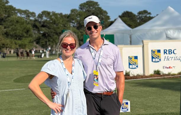 Madison and Jack wear business casual, sunglasses, and lanyards, and stand outside on the green for a golf tournament. 