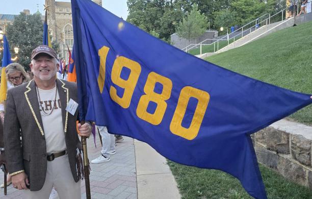 Mark holds the Class of 1980's blue and yellow flag and smiles. He wears a Lehigh t-shirt and hat.