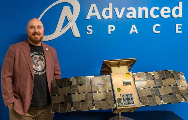 Sean Hoenig stands alongside a model of the Capstone spacecraft and the Advanced Space logo.