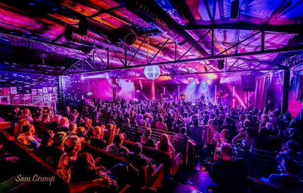 The State Room mid-concert. Band members play on stage, the crowd watches, and purple light fills the room.