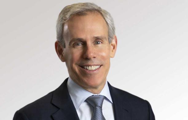 John wears a dark suit jacket, light shirt, and a tie. He smiles. 