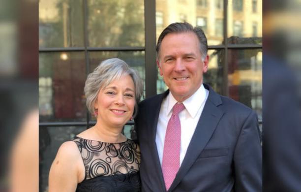 Dressed in semi formal attire, Ann and Weston Hicks pose smiling for a photographer in front of a wooden door with windows.