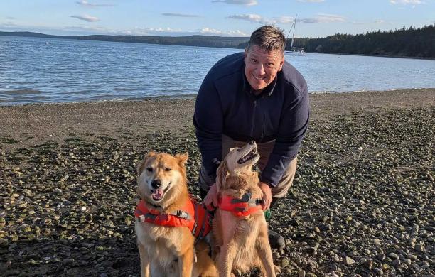 Mark bends down and pets two dogs on a pebble beach with clear water and trees visible behind him.