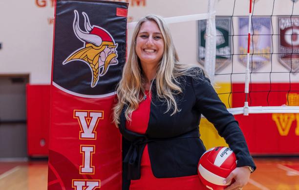 Rochelle Sheridan poses holding a red and white volleyball in a red dress and black blazer on an indoor volleyball court beside the pads protecting the net posts, reading “Vikings.”