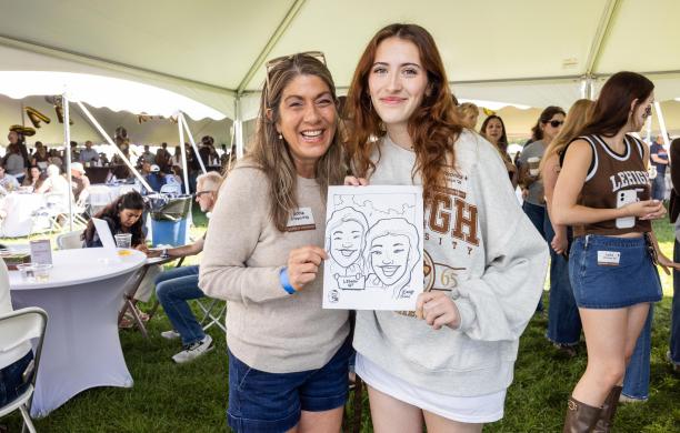 A student and their parent stand under a tent and smile while holding up a caricature of them taken at the event. 