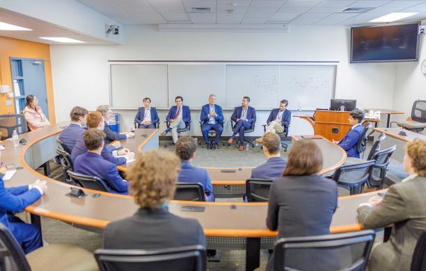 Five alumni wear professional attire and sit facing a room full of attendees. The crowd is also dressed in business attire.