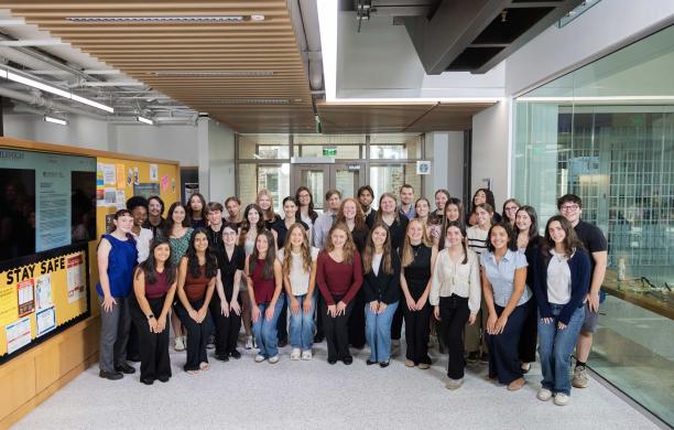 A large group of Soaring Together Scholars pose together in a lobby. They all wear business casual and smile. 