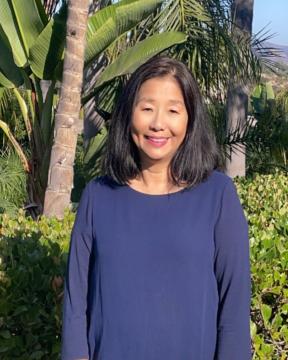 A woman with brown hair and wearing a blue sweater smiles in front of palm trees on a sunny day.