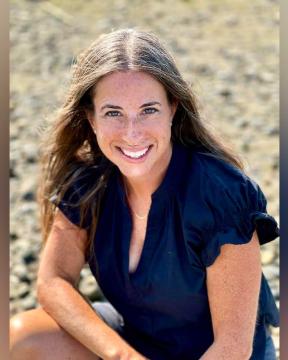 A woman with brown hair and a black top smiles at the camera while sitting outdoors.