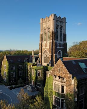 A tall, historic stone building with a central tower and ivy-covered walls, surrounded by trees under a clear blue sky.