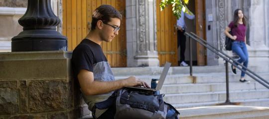 Student sitting on the steps of Alumni Memorial with his laptop