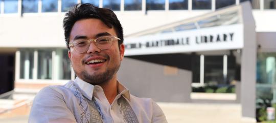 Adrian Suarez ’22 ’23G sitting in front of the E.W. Fairchild-Martindale Library
