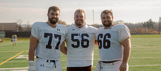 Three football players (numbers 77, 56, and 66) stand shoulder-to-shoulder on the field in a friendly camera-facing pose.