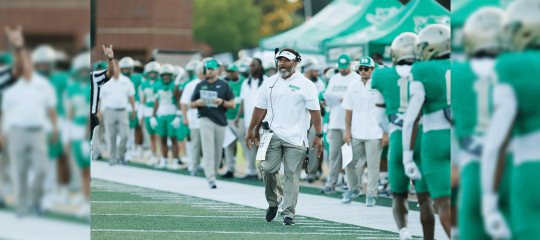 Coach Bryant Appling walks down the field, calling plays to his football team with Buford players beside him on the sidelines.