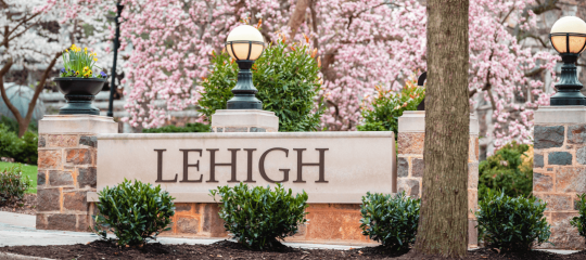 A stone bench with the word Lehigh etched across the front and beautiful cherry blossoms and trees in the foreground