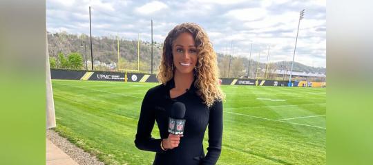 Kayla Burton holding a microphone and standing before a yellow goalpost on a lush, green football field.