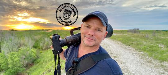 Alumni Owned Business, Michael Patrick O'Neill smiling for a selfie with his camera with an expansive field behind him.
