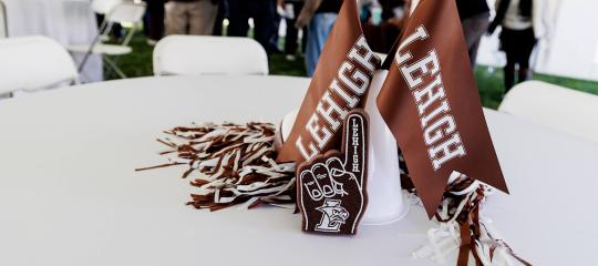 Brown Lehigh flags, pompoms, ad a small brown foam finger sit on a table arranged among others under a large white party tent.