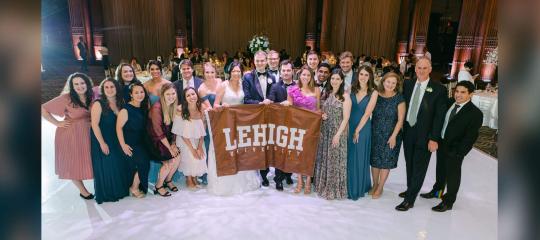 A large group of people surrounding Becky Lok and Mike Meehan at their wedding reception, holding a Lehigh flag.