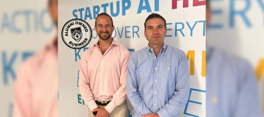 Business partners wearing button down shirts stand smiling in front of a wall emblazoned with brightly colored words.