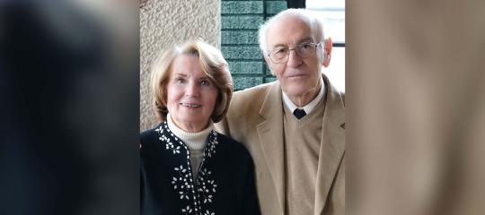 Sally Gordon stands to the left of her husband Michael in 2025, as he holds her right hand with his right hand.