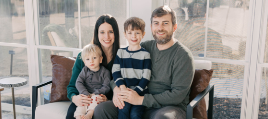 Family sits for a portait in a well lit white room against a wall of windows, with father and mother each holding a small boy in their lap.