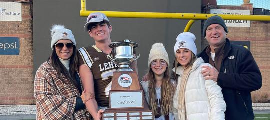 A Lehigh football player holding a large championship trophy stands on the field with four smiling family members.