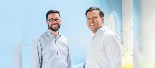 In a brightly lit room with baby blue walls, two men stand casually facing each other and smiling for the camera while wearing business shirts with no ties.