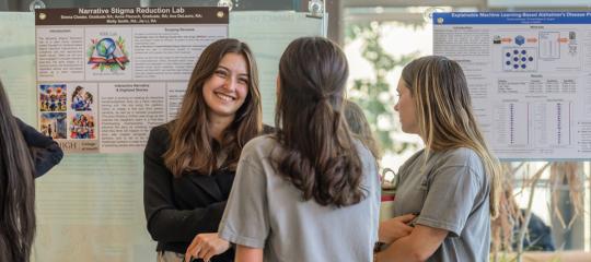 A smiling student discusses research posters, including "Narrative Stigma Reduction Lab," with peers at a College of Health symposium at Lehigh University.