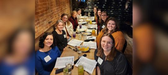 A group of women sit at a long dining table with menus in front of them, posing for the camera.
