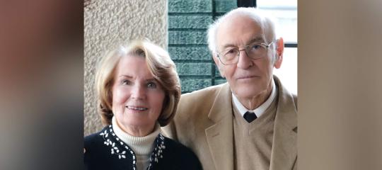 Sally Gordon stands to the left of her husband Michael in 2025, as he holds her right hand with his right hand.
