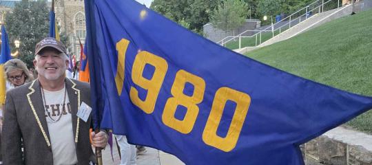 Mark holds the Class of 1980's blue and yellow flag and smiles. He wears a Lehigh t-shirt and hat.