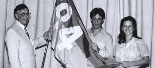 A black and white photo of a man in a light colored suit standing with two students on stage, all holding a large flag together that reads “’84.”