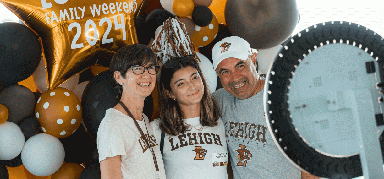 A family of three smiles for a photo while standing infront of brown and white balloons.
